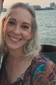 A man with dark gray hair in a shirt and blazer stands on the left of a shorter woman with blonde hair in a patterned top. They are smiling and a river with land on the other side is visible behind them.