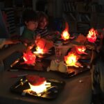 Photograph of a dark room with a bunch of lit-up campfire robots on a tabletop with kids sitting nearby