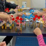 5 completed proppy jalopy robots with googly eyes and magic marker designs and children's hands in the frame adjusting the robots.