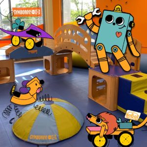 Colorful photograph of a Gymboree playspace with illustrated Brooklyn Robot Foundry mascots superimposed. PetPuppyborg robot climbing on soft dome; BabyBambino (lion) sculpture rolling in front, BitsyBot character standing on play structure; Proppy Jalopy flying in from side.