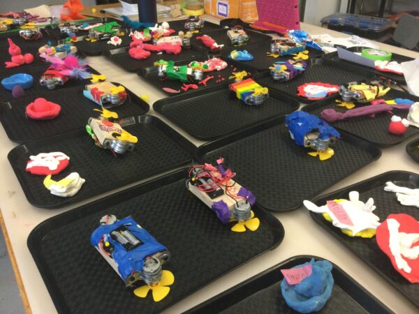 Photo of a table hold many trays with kid made robots on them.