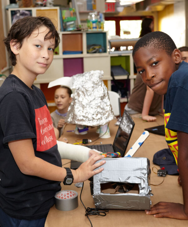 Two kids look to camera , taking a pause from working on a robot project together on the table