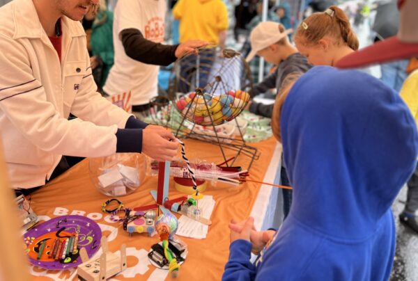 A colorful table with a robot that makes pipe-cleaner bracelets and a spinning bingo wheel of balls in the background.