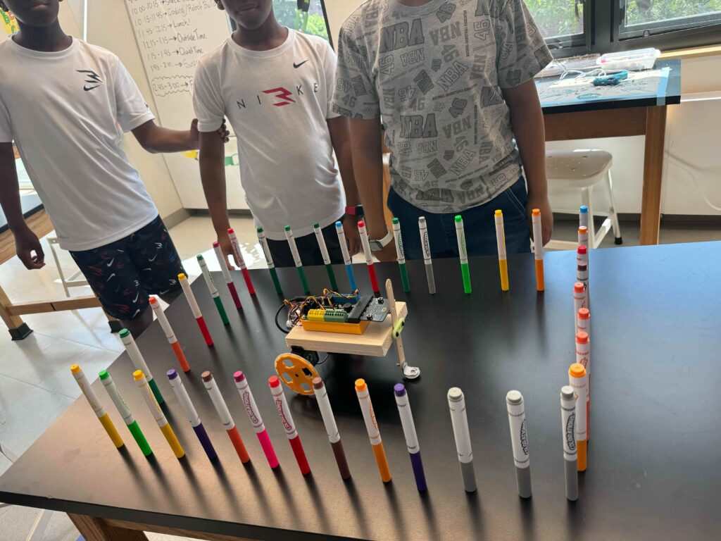 3 students stand near a table with a robot on it, surrounded by approximately 40 markers all standing up around it in a rectangle