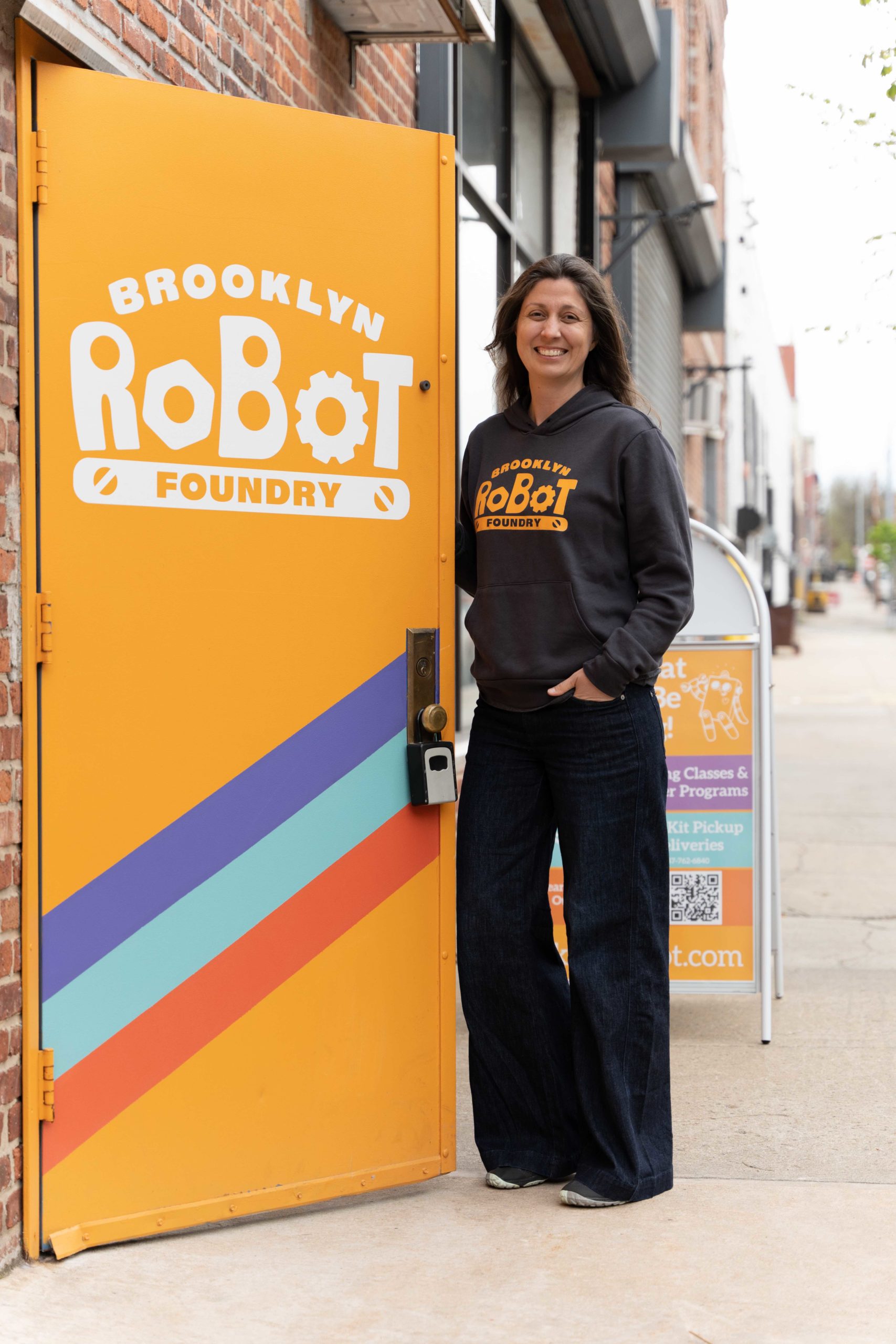 Profile picture (full body) of Jenny Young. She is wearing a grey Brooklyn Robot Foundry sweatshirt and dark pants, standing outside of the orange Brooklyn Robot Foundry door. The blurred city block is behind.