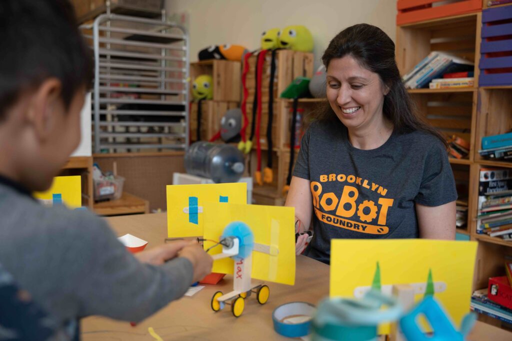 Jenny Young, owner of Brooklyn Robot Foundry, smiling as she sits at a table playing with robots with a child (of whom we see only a portion of their side).