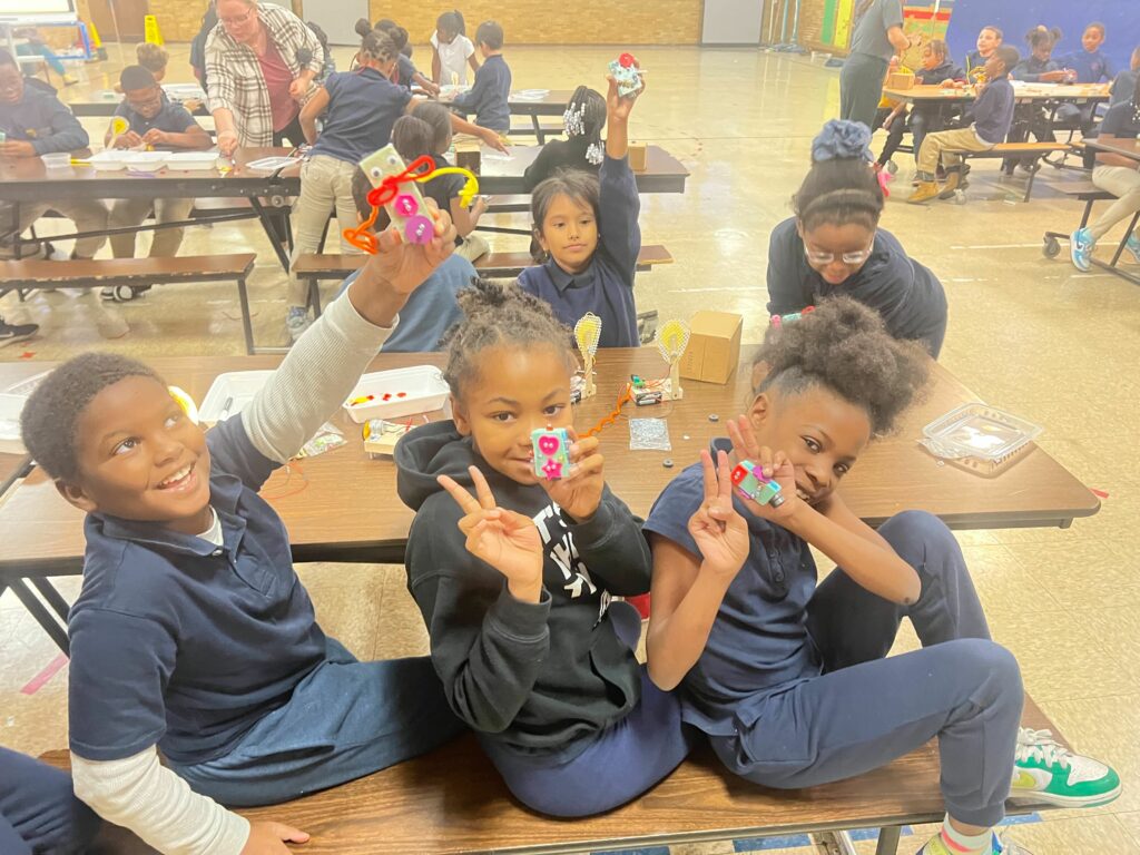 A group of kids sitting at a cafeteria table proudly holding up the little bots that they built.