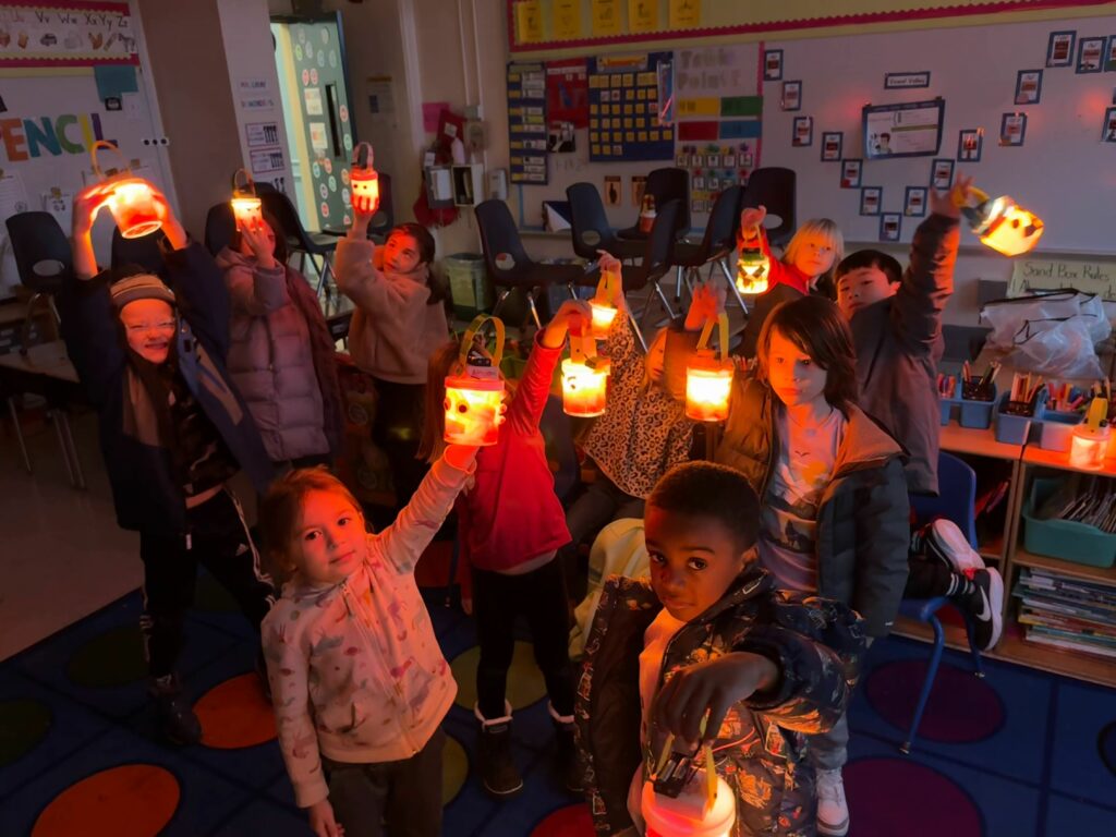 A classroom of kids in the dark, holding up their glowing robots and smiling.