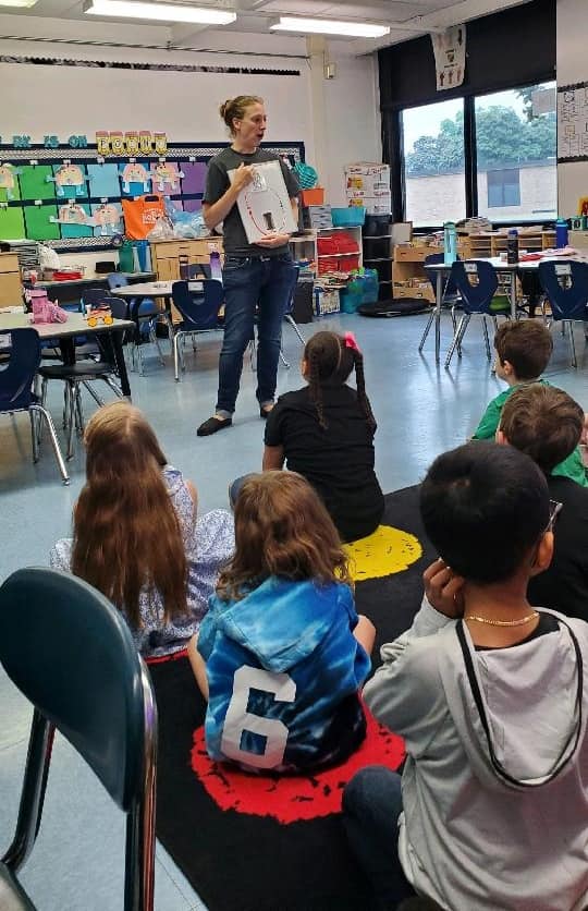 Teacher standing in front of a classroom of kids sitting on the floor. The teacher is holding up a display of a circuit.