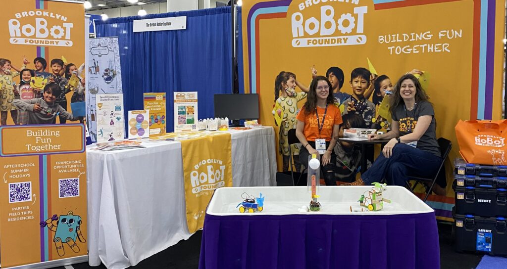 Photograph of Jenny and Tawny sitting at a table in the Brooklyn Robot Foundry booth at IFE