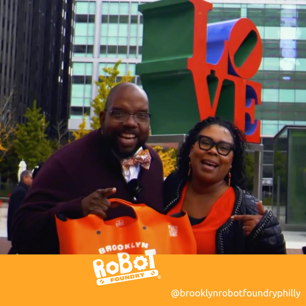 Denise Marie and Richard Snow stand in front of Philadelphia's LOVE sculpture while holding an orange Brooklyn Robot Foundry bag. The Brooklyn Robot Foundry logo next to "@brooklynrobotfoundryphilly"